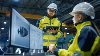 Two persons are looking at schematics on a computer in an industrial setting.
