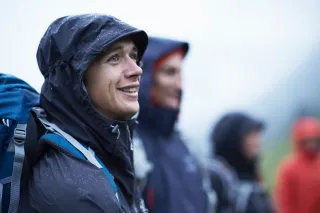 People in weather resistant clothes are standing in the rain and the man in the front is smiling